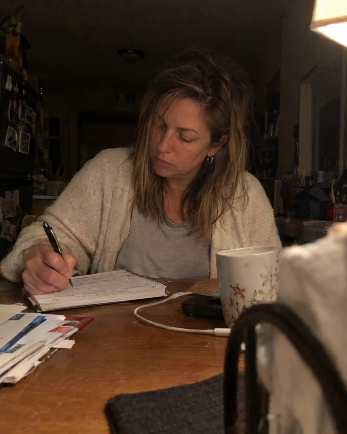 Woman writing in a notebook alone at her kitchen table late at night, phone propped against a napkin holder