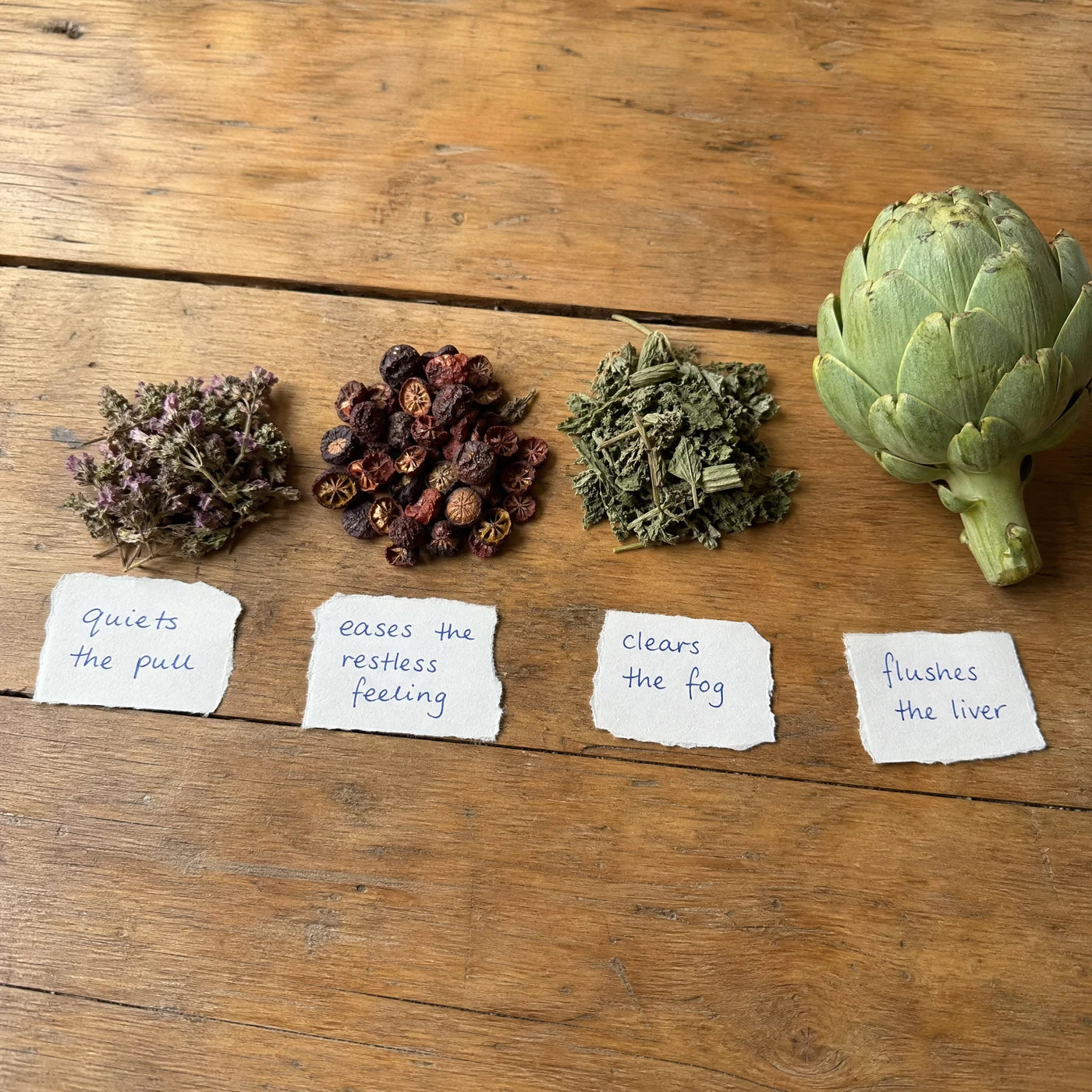 Four plant ingredients laid out on a wooden table, each labeled with its job: quiets the pull, eases the restless feeling, clears the fog, flushes the liver