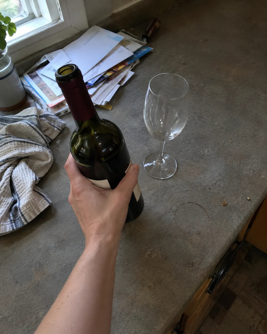 First-person view of a hand reaching for a wine bottle and empty glass on a kitchen counter in early evening light