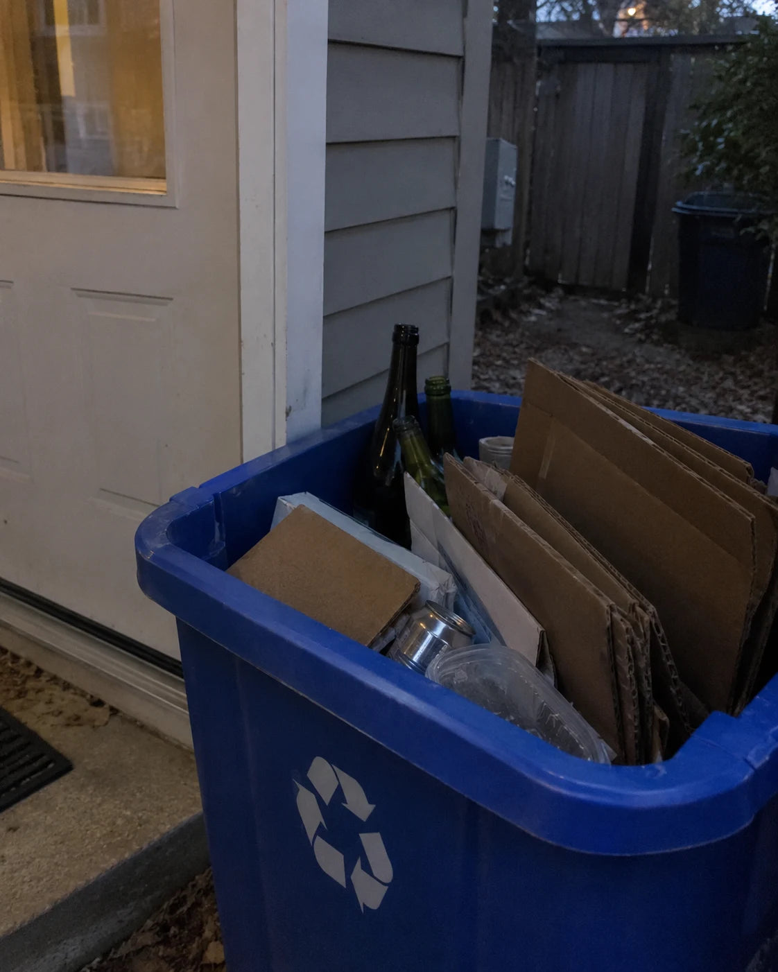 Empty wine bottles tucked toward the back of a home recycling bin near a back door at dusk