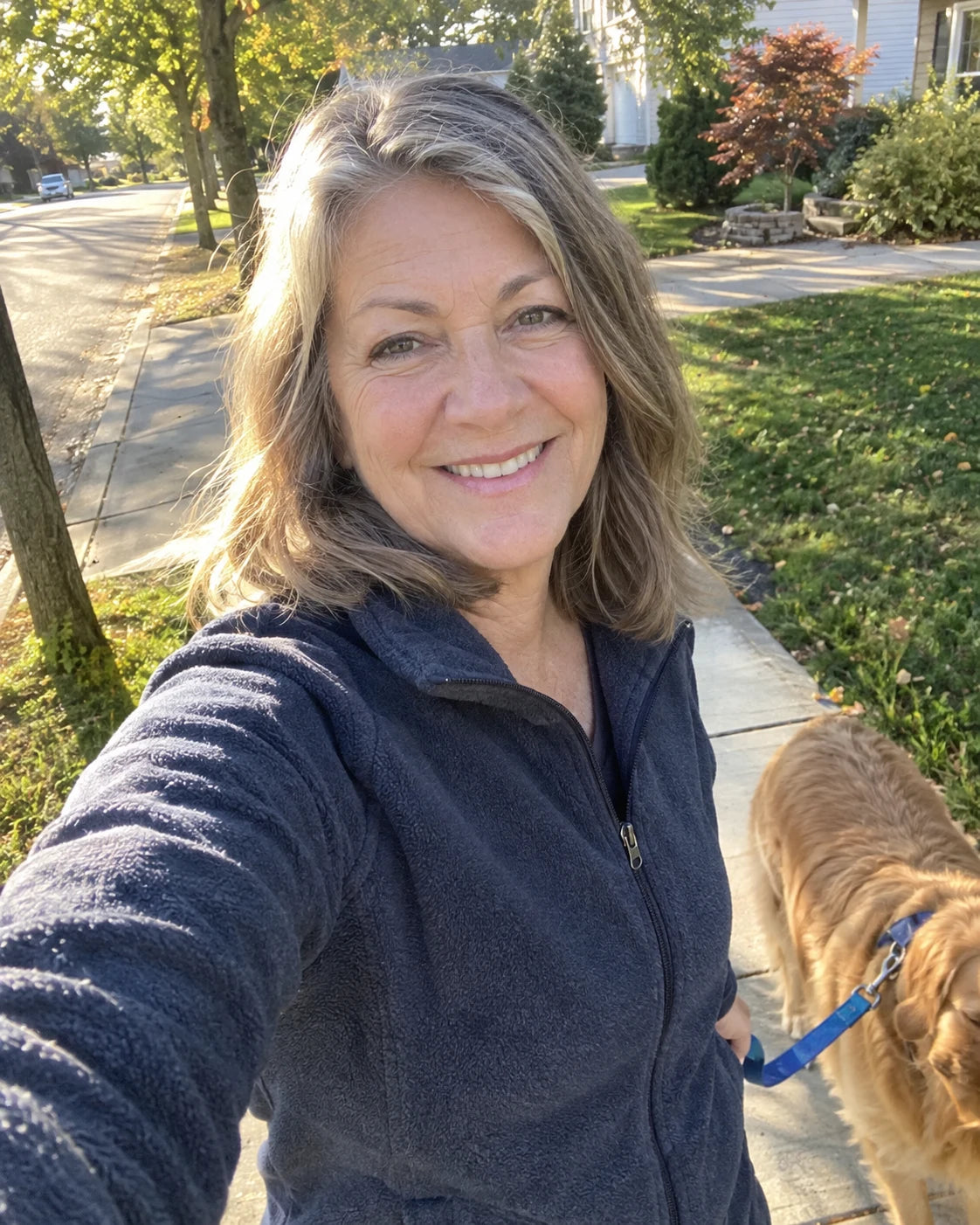 Woman in her 60s taking a selfie with her golden retriever while walking him in the early morning