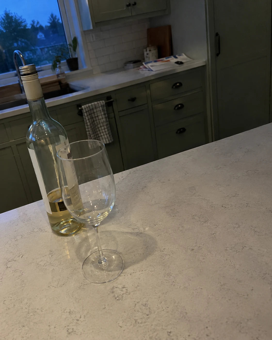 An open bottle of white wine and an empty glass sitting on a kitchen counter at dusk