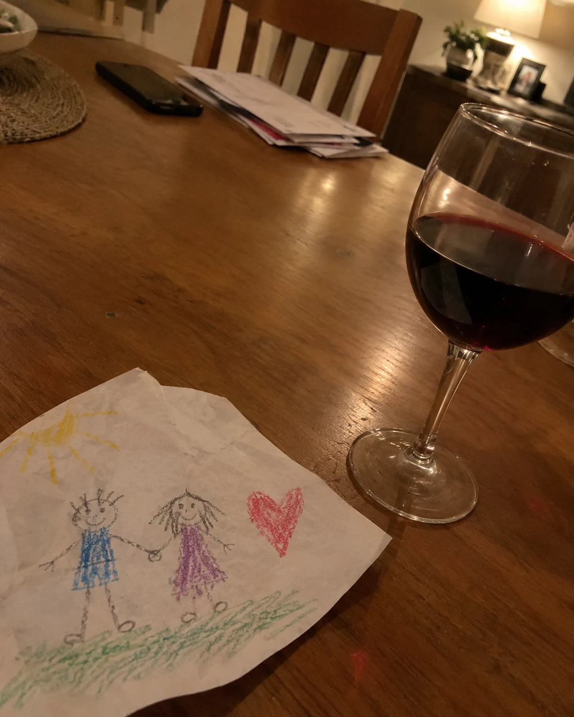 A glass of red wine on a dining table next to a child's crayon drawing of two people holding hands and a heart