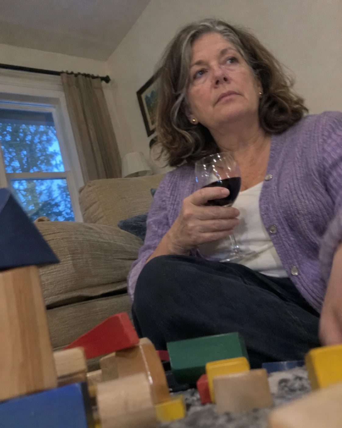 Grandmother sitting on the couch with a glass of red wine, looking distant, grandkids' building blocks out of focus in the foreground