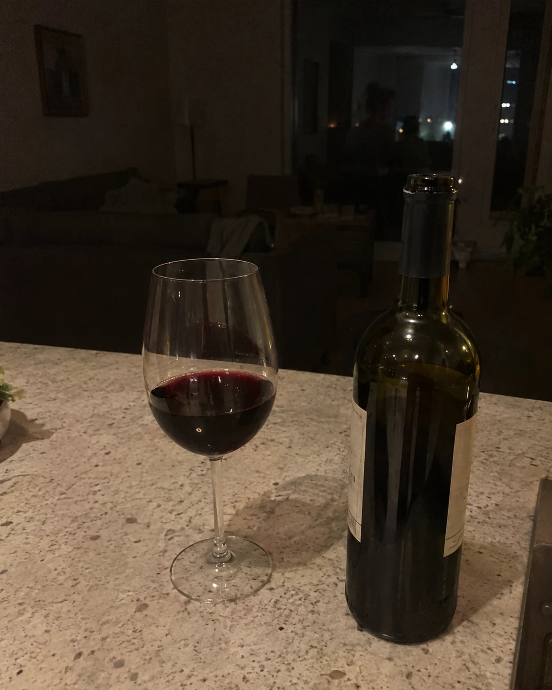 A glass of red wine and an open bottle on a dark kitchen counter at night