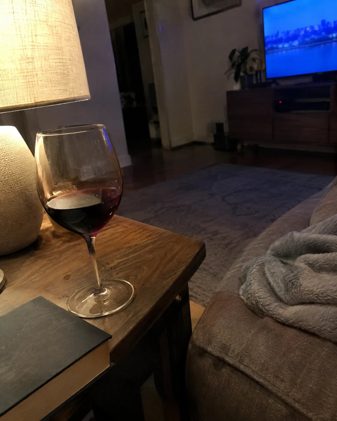 A poured glass of wine on a side table next to an unopened book and a muted TV in the background