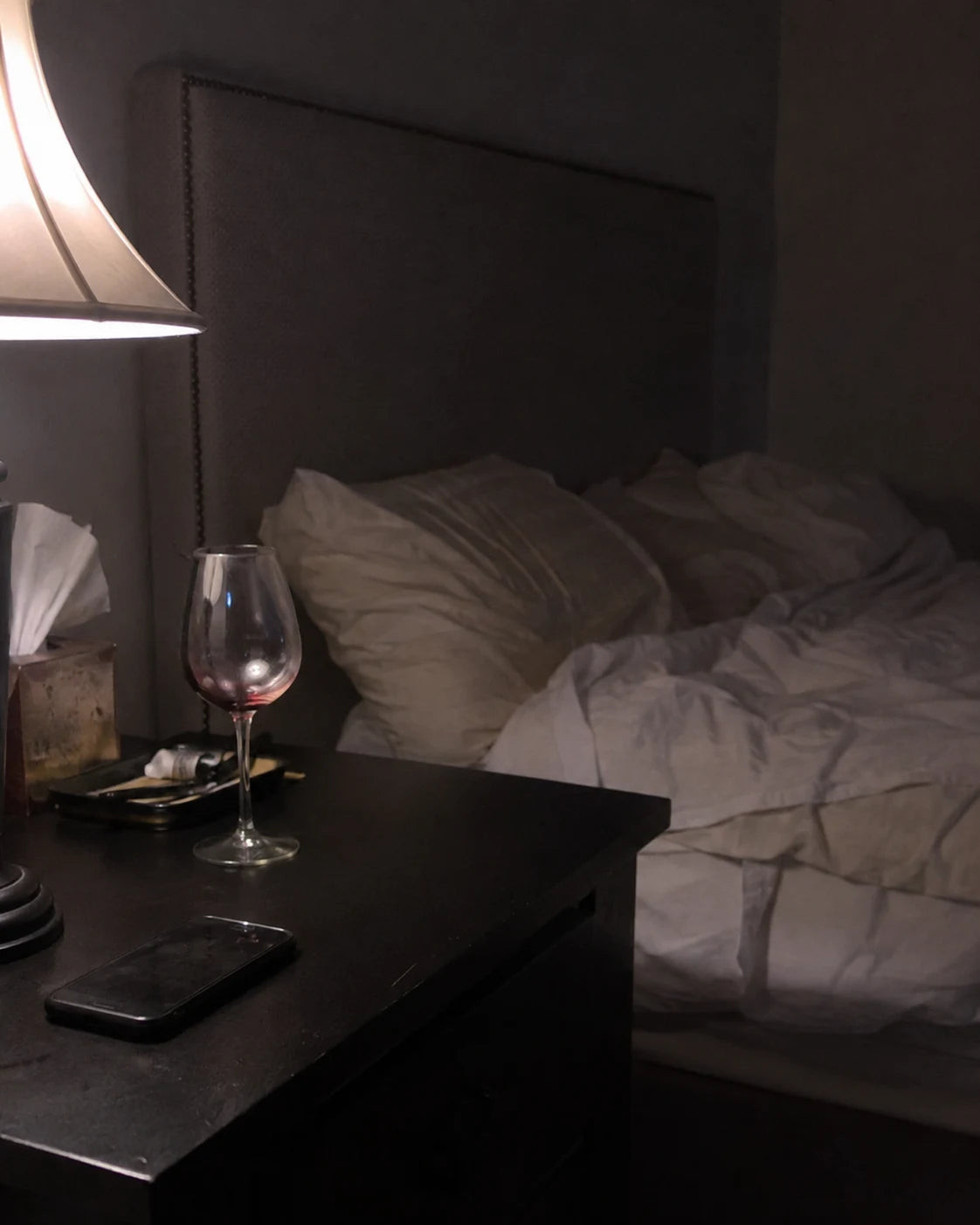 A mostly-empty wine glass on a bedroom nightstand next to a lamp and a phone, bed in the background