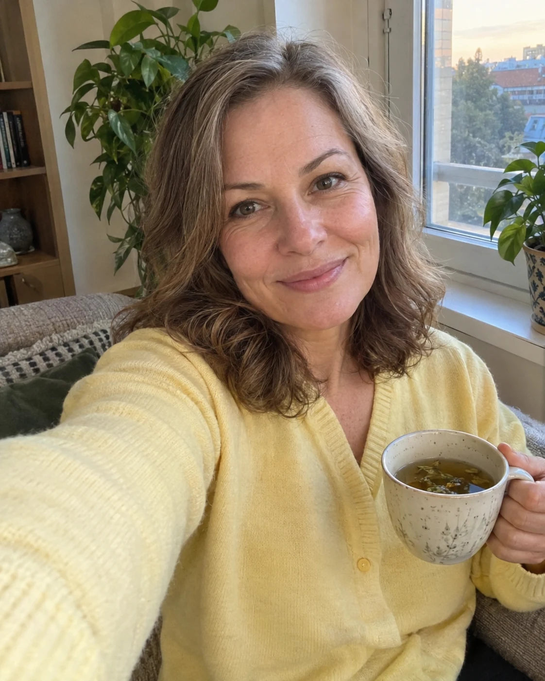 Woman smiling and relaxed by a sunlit window holding a cup of tea