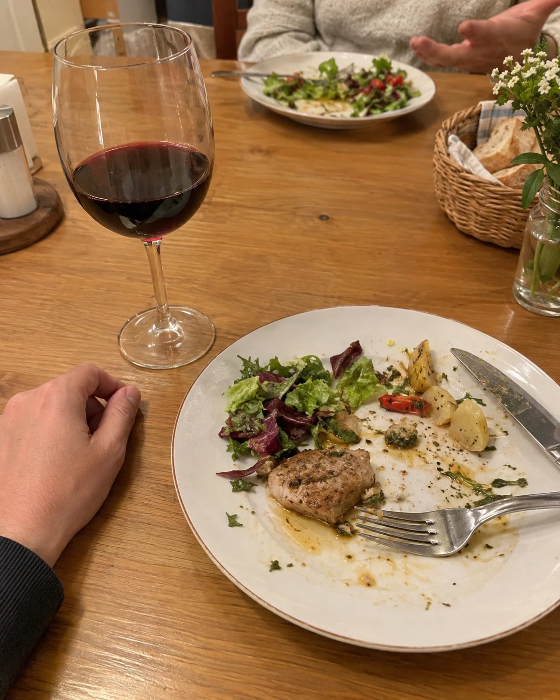 Point of view at a dinner table, wine glass and half-eaten plate, a hand resting nearby while someone across the table talks