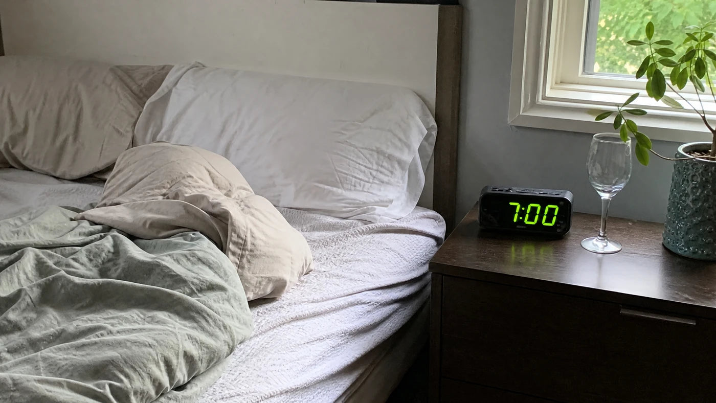 A rumpled bed in soft morning light with a nightstand clock showing 7:00 and an empty wine glass